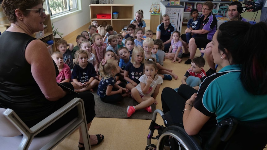 A woman in a wheelchair and another woman sit in front of a group of preschool students, reading a story.