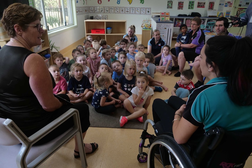 A woman in a wheelchair and another woman sit in front of a group of preschool students, reading a story.