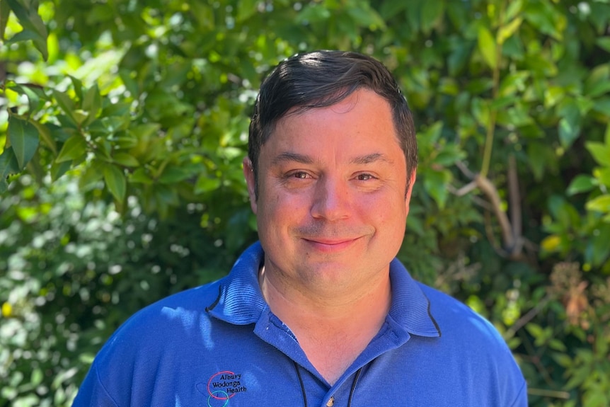 A man smiles at the camera in a blue polo top with an Albury Wodonga Health logo on the right breast in front of some greenery