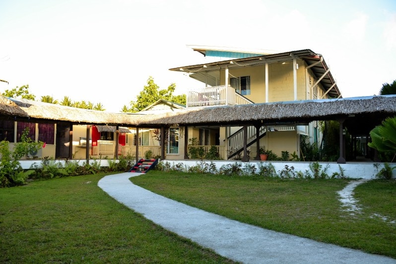 A path leading through green grass to a yellow building with a thatch roof.