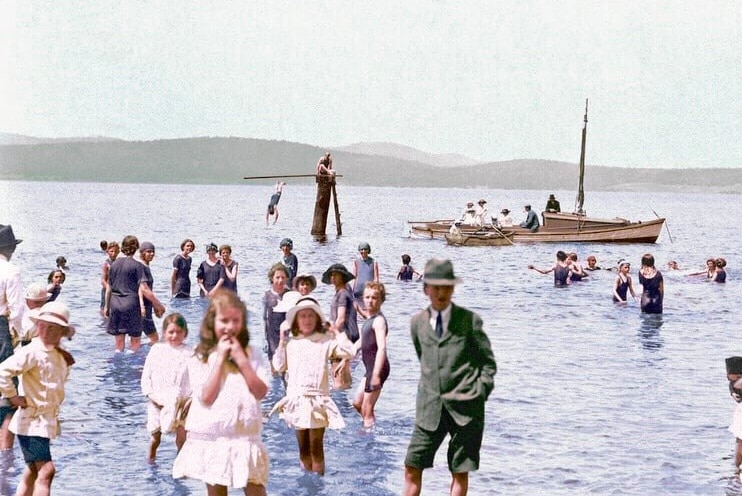 Children and adults frolic in the shallows, people in the background row a boat and others dive off a pole.