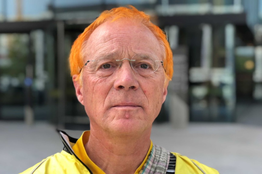 A man with orange hair and glasses looks serious outside a building.