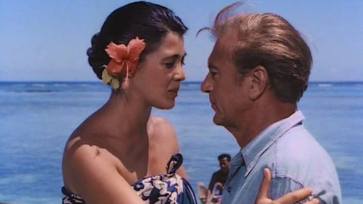 A woman wearing flower in ear and ie lava lava looks into eyes of man placing her right hand on his shoulder. Blue ocean behind 