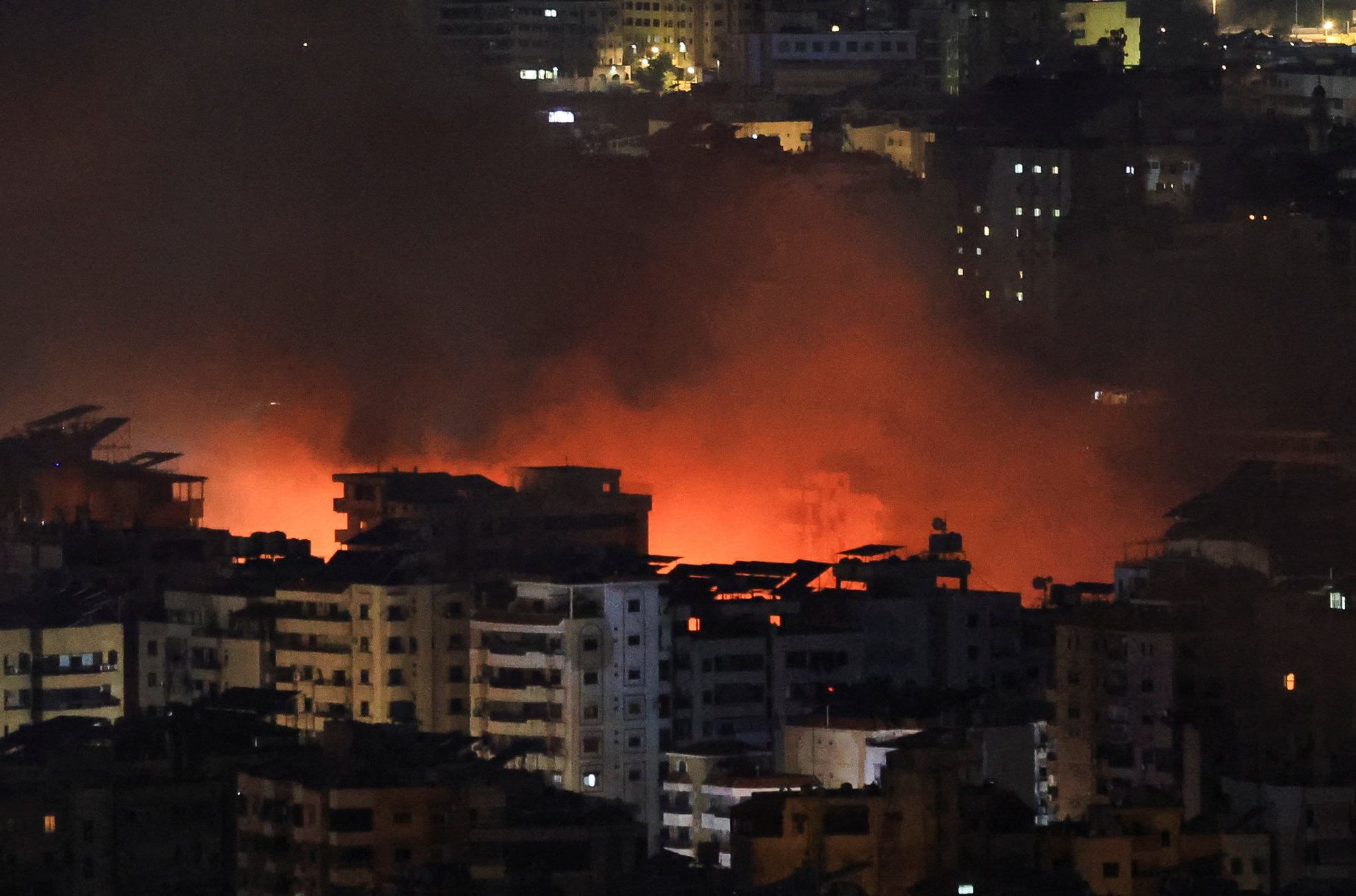 Orange smoke rises between buildings on a city skyline at night