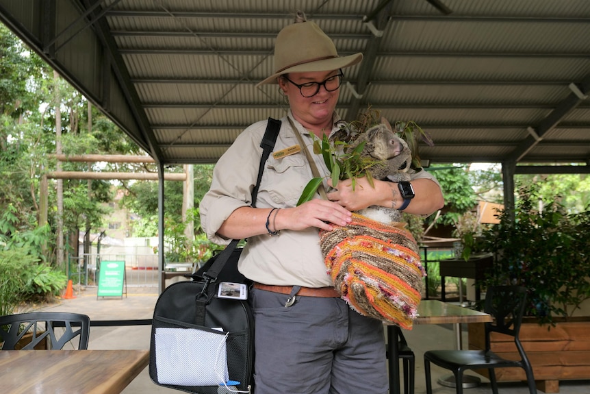 Woman with broad-brimmed hat, carrying baby koala in orange knitted bag and black bag on other shoulder