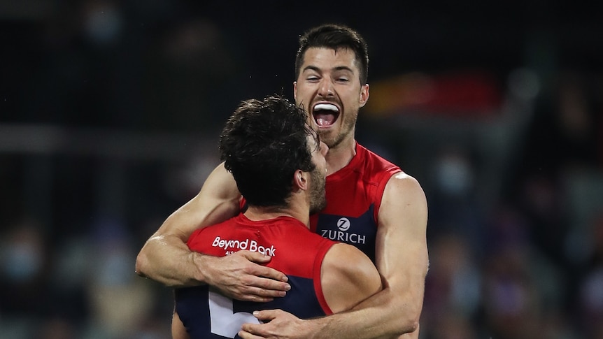Two Melbourne Demons players celebrate a goal by hugging and jumping into their air during an AFL game.