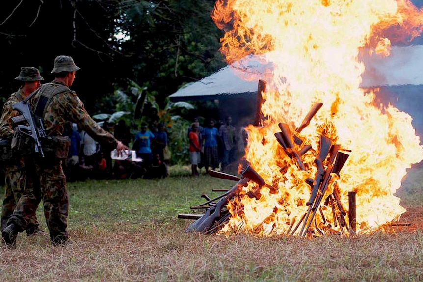 A burning pile of guns with men in army uniform looking on.