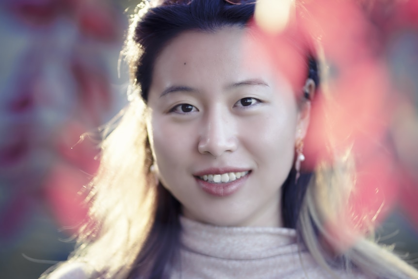 A picture of a woman of Chinese heritage smiling with blurry red leaves around her face. 