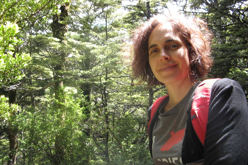 A woman with dark curly hair smiles as she hikes through lush forest.