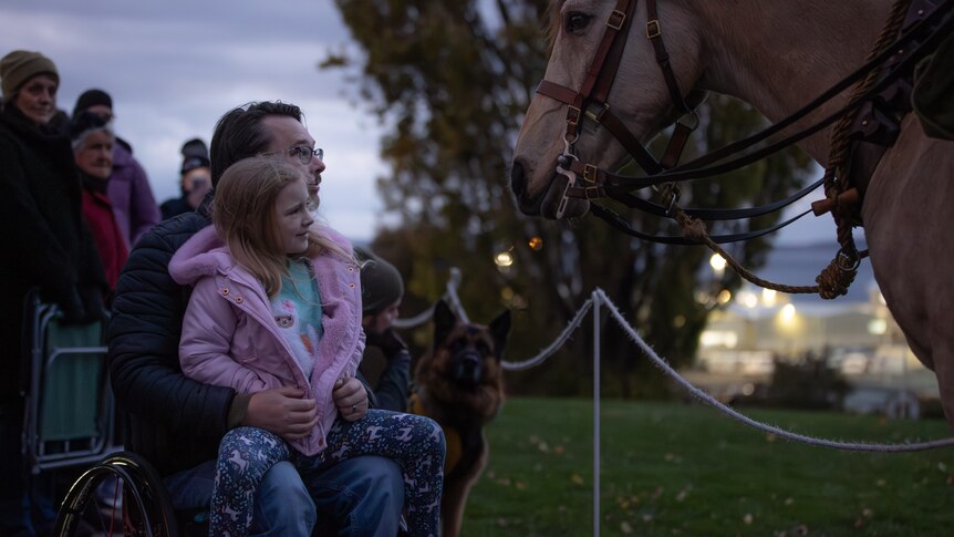 A man in a wheelchair holds his young daughter on his lap as they look a horse in the force.