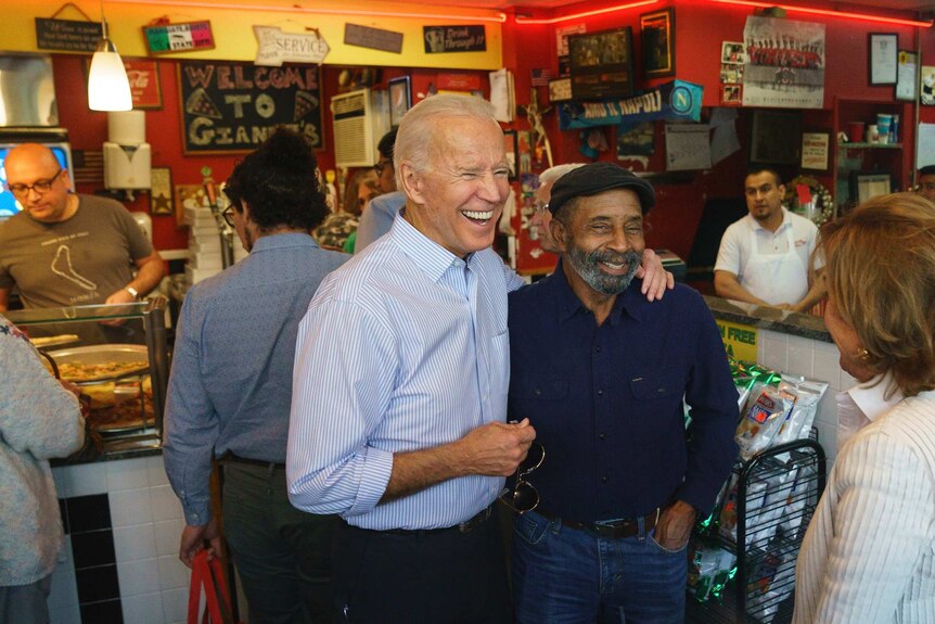 Joe Biden smiles and stands with his arm around a man wearing a black beret.