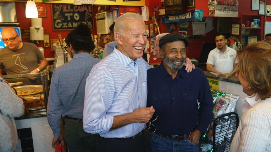 Joe Biden smiles and stands with his arm around a man wearing a black beret.
