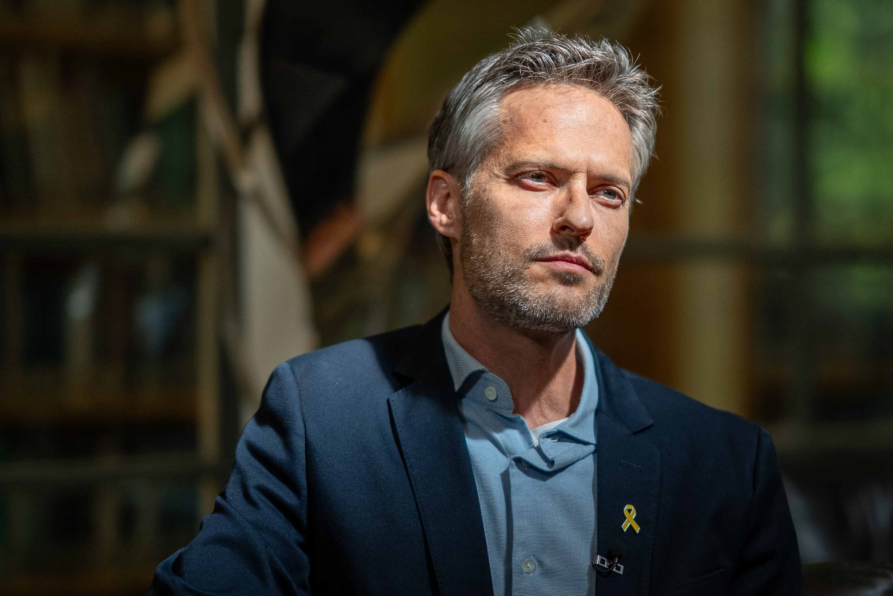 A man with grey hair looking up and to the right, in sunlight, on a dark backdrop.