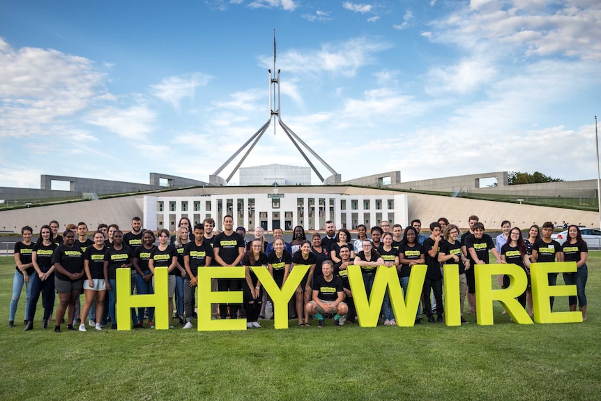 Large group of Heywire winners standing on grass with Parliament House in background.