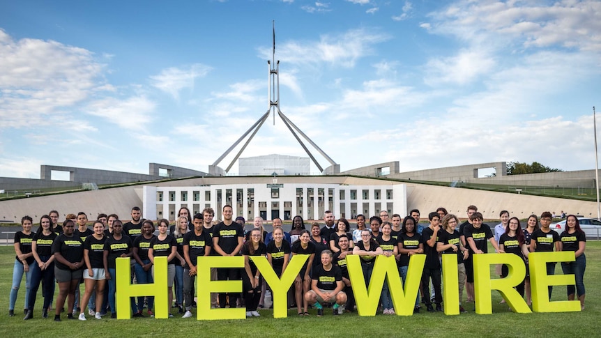 Large group of Heywire winners standing on grass with Parliament House in background.