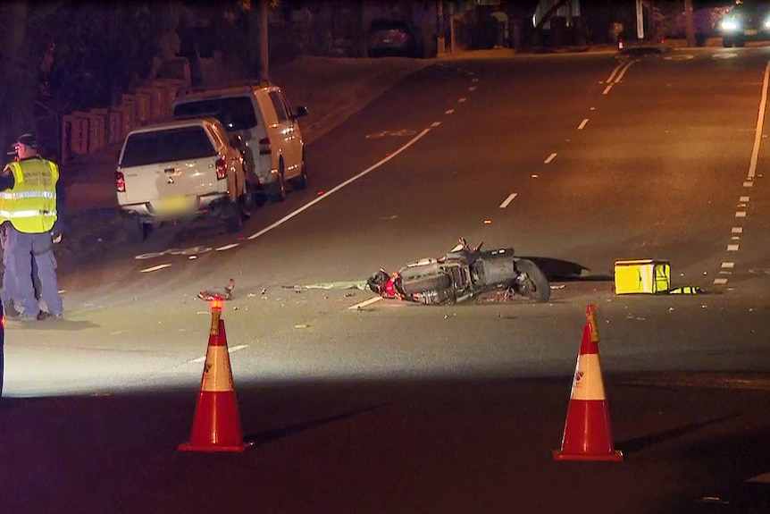 police at a crash scne stand on the side of a road as a smashed up motorcycle lays on the road