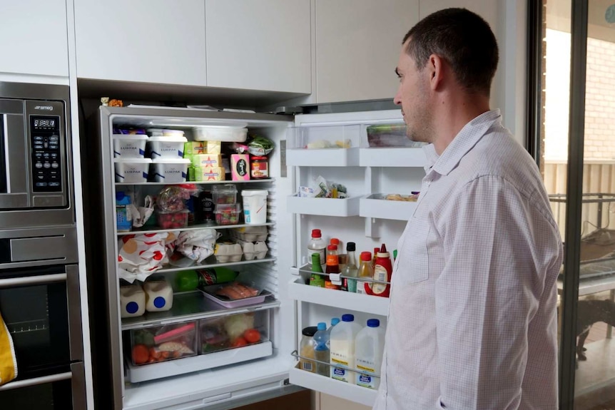 A man in a white business shirt regards the contents of his large, well-stocked fridge.