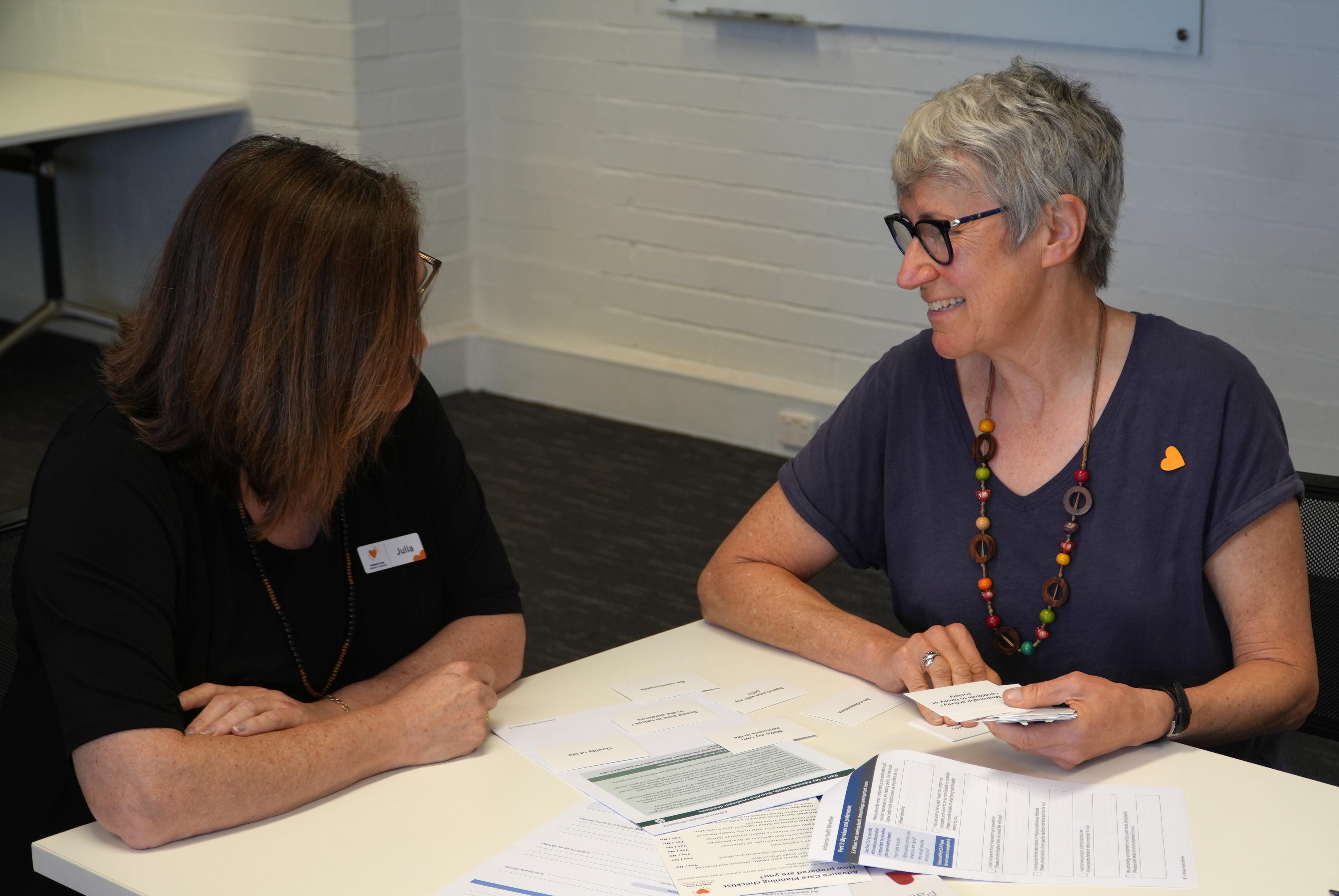Duas mulheres estão sentadas à mesa olhando papéis. 