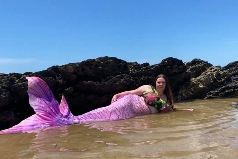 Une femme allongée sur la plage portant une queue de sirène rose.