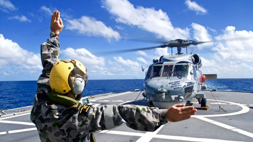 The Seahawk is on Arunta's deck, at sea. A Navy member is in the foreground signalling.