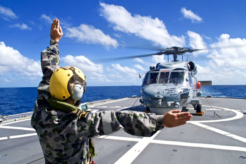 The Seahawk is on Arunta's deck, at sea. A Navy member is in the foreground signalling.