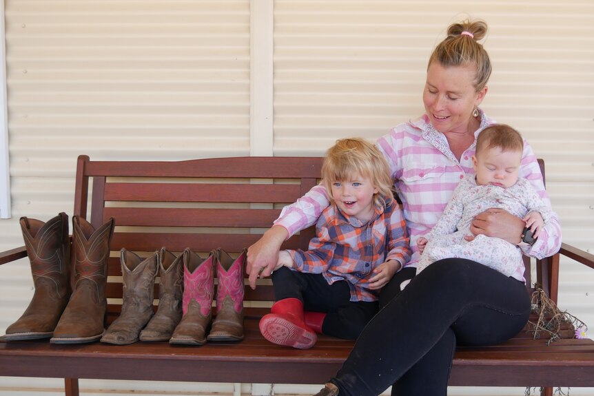 A woman, child and infant wearing farm clothes sit on a bench alongside with western boots. The woman smiles at the child.