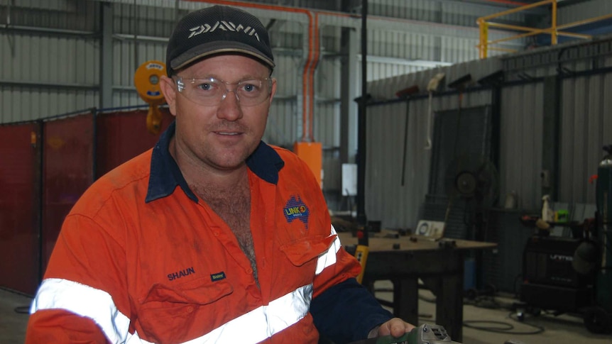 A man in a high-visibility fluoro long-sleeved shirt, and work safety glasses, stands in an industrial-type shed.