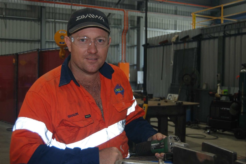 A man in a high-visibility fluoro long-sleeved shirt, and work safety glasses, stands in an industrial-type shed.