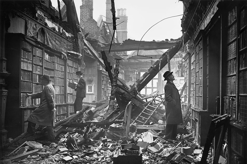 Three civilians still looking at books in a London library with rubble around them and the roof destroyed by a bomb.