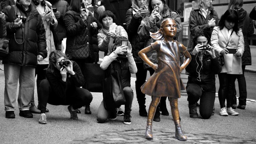 A bronze statue of a fearless young girl with her hands on hips. A crowd of people behind the statue are taking photos of it.