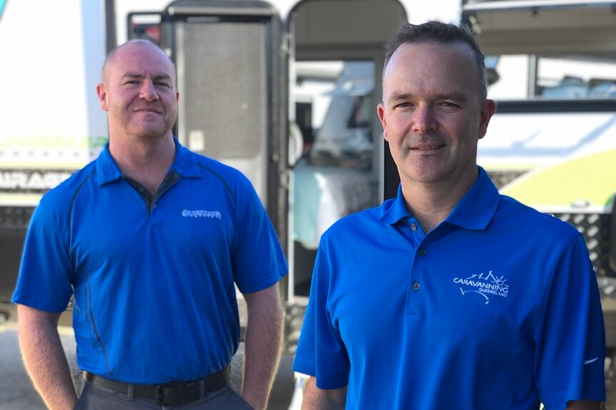 Two men in blue shirts smiling in front of a caravan