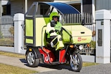 A postie clad in fluro yellow clothes rides a motorised three wheeled motorbike which has a sunshade over the top.