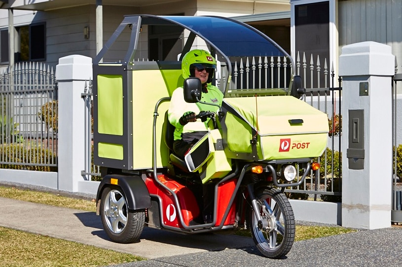 Un postie vestido con ropa amarilla fluro monta una motocicleta motorizada de tres ruedas que tiene una sombrilla en la parte superior.