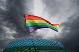 A rainbow flag flutters in the wind with dark grey skies in the background and a lit-up stadium.