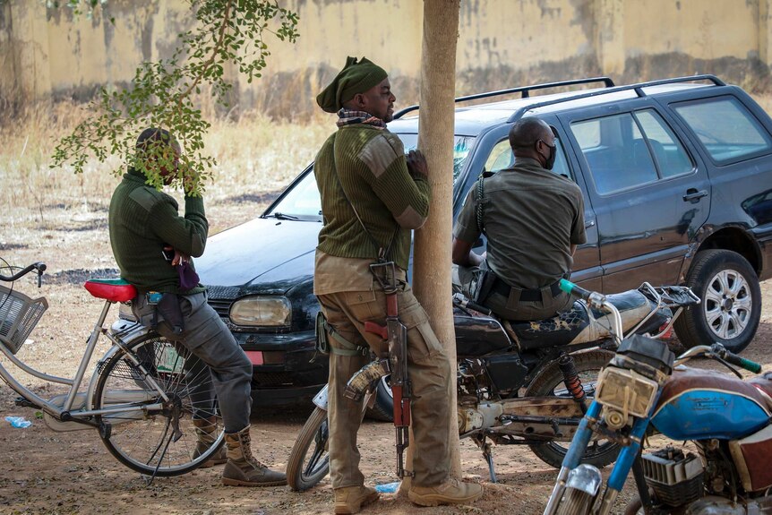 Three armed security guards, one sitting on a bike, one standing and the other sitting on a motorbike look in the same direction