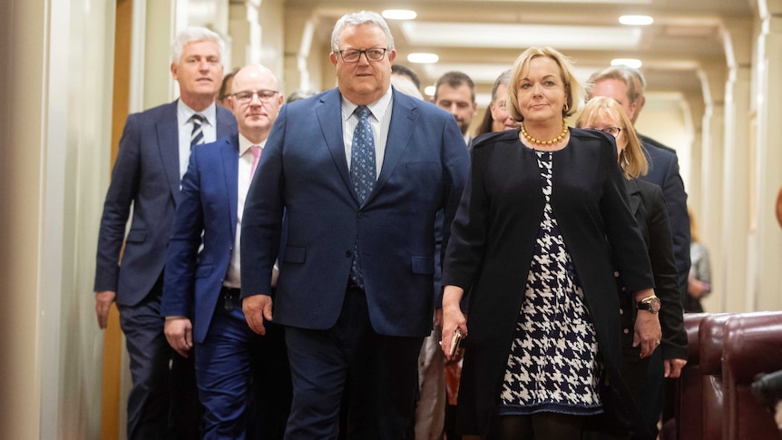 A group of middle aged people walk down a hallway, lead by a woman in a blue jacket