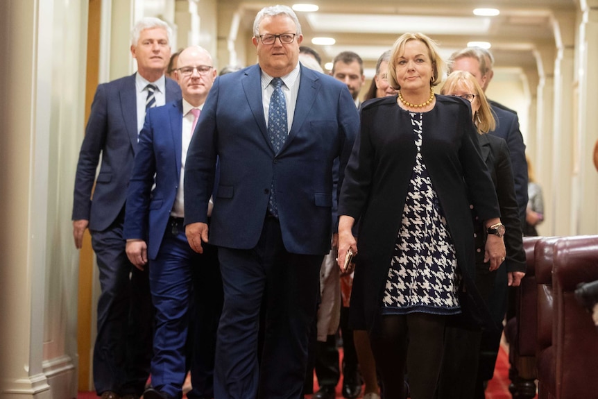 A group of middle aged people walk down a hallway, lead by a woman in a blue jacket