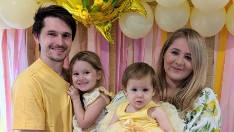 Two parents holding a child each stand in front of yellow and pink ribbons, everyone smiling.