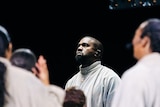 A choir stands around a well-lit Kanye, who looks to the sky