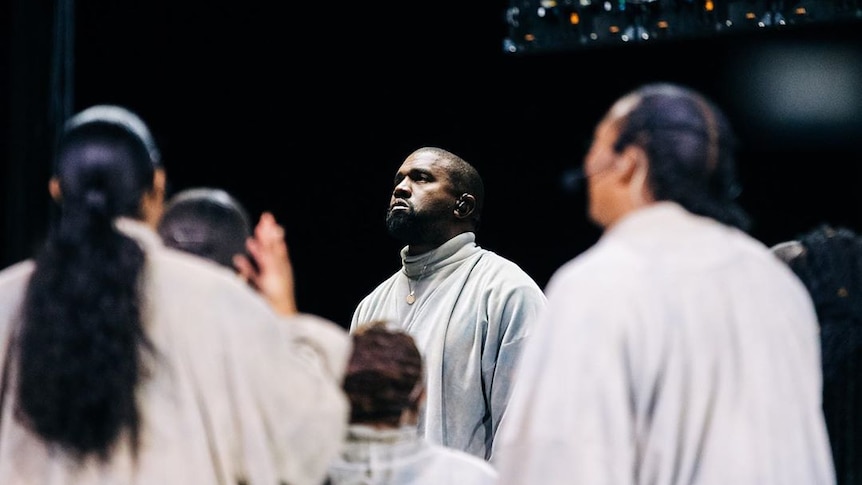 A choir stands around a well-lit Kanye, who looks to the sky