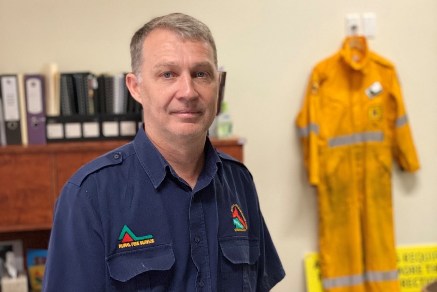 Man in navy blue rural fire shirt sitting on a desk in an office with a rural fire service uniform hanging in the background