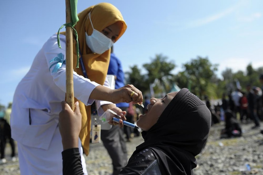 A female healthcare worker administers an oral vaccine to a girl wearing a head covering