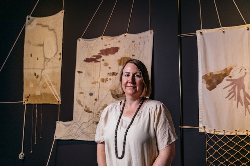 White woman with should-length blonde hair wears cream shirt and black necklace in front of fabric artwork hung on gallery wall.
