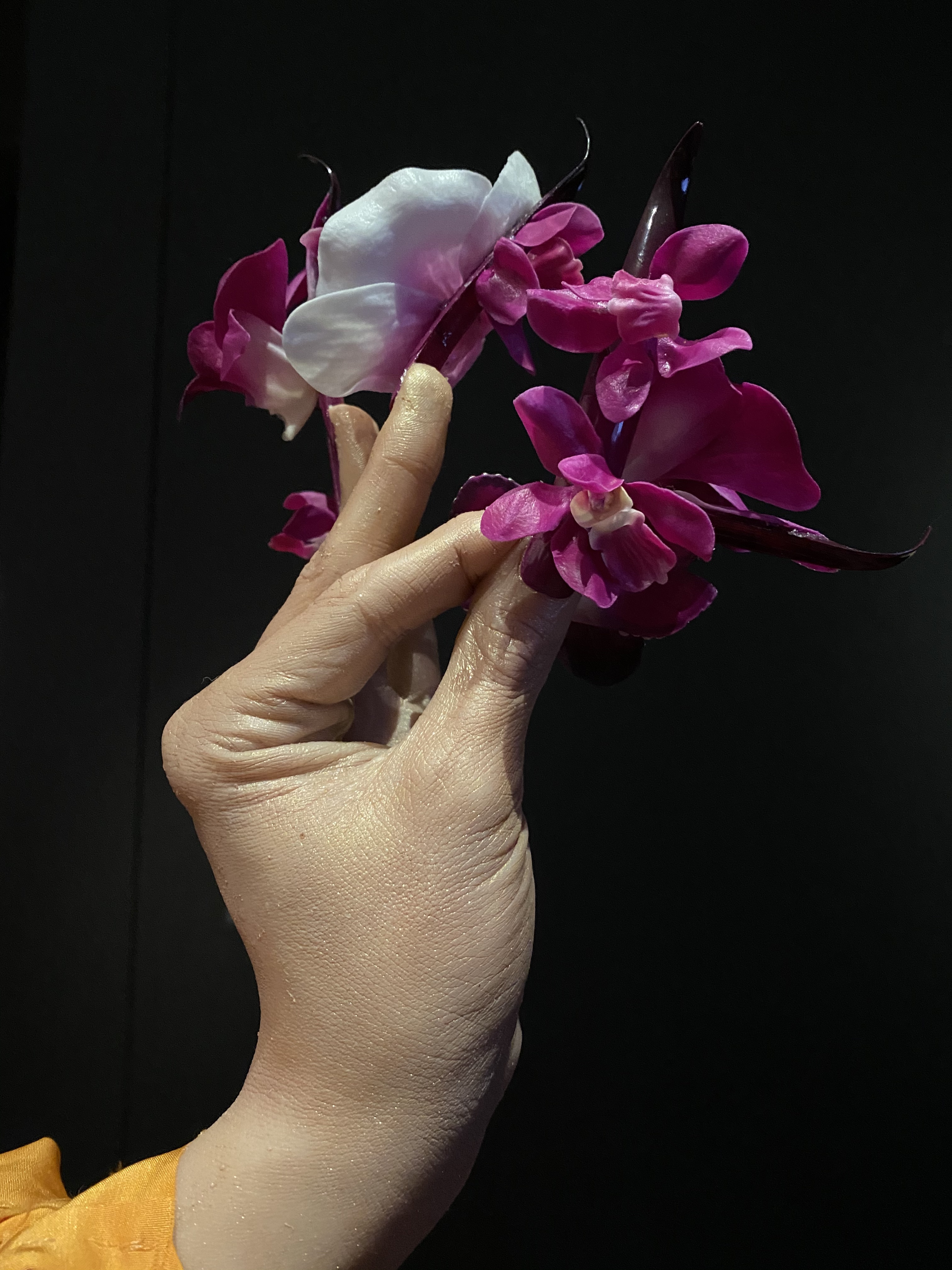 DETAIL from a sculpture, showing a hand with long purple acrylic nails holding orchids.