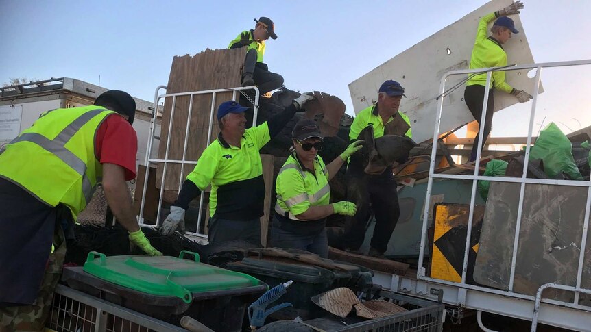 Six volunteers wearing caps and green high-visibility shirts put rubbish into a trailer.