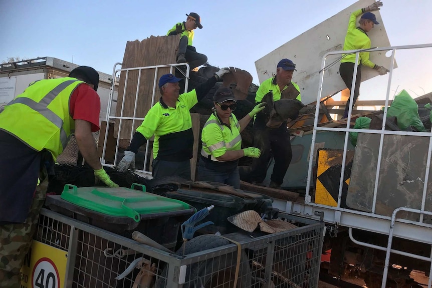 Six volunteers wearing caps and green high-visibility shirts put rubbish into a trailer.