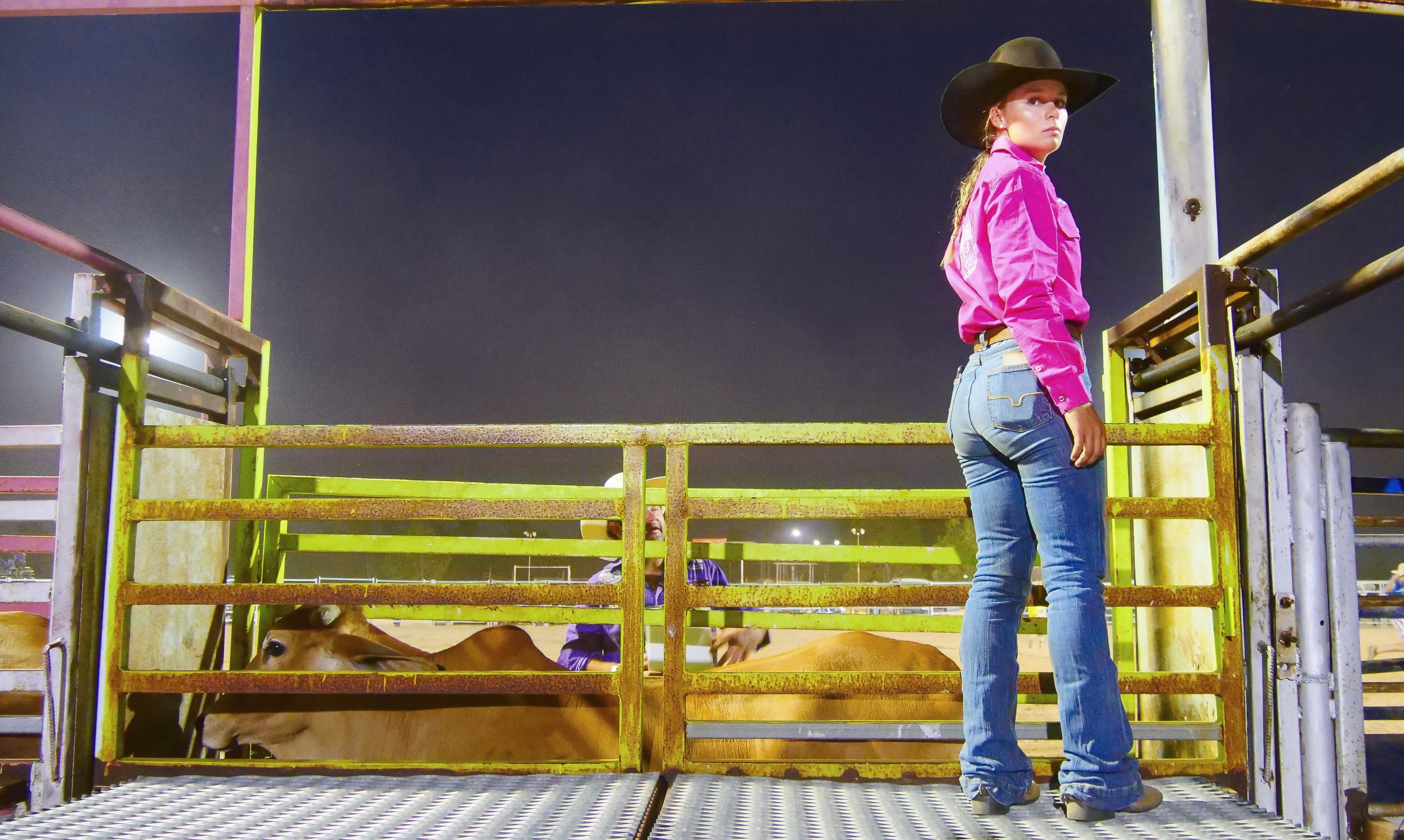 A cowgirl wearing blue jeans and a pink shirt stands behind chutes a a rodeo
