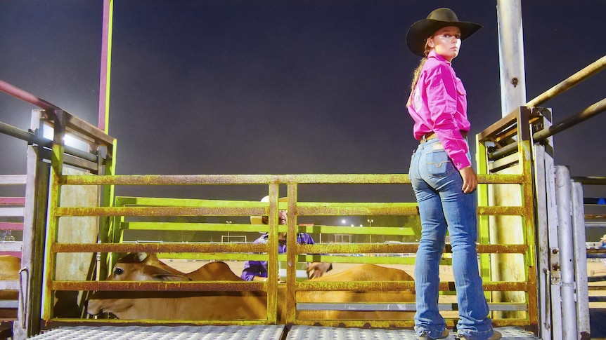 A cowgirl wearing blue jeans and a pink shirt stands behind chutes a a rodeo