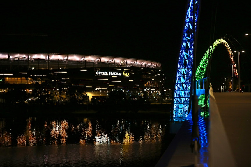 Perth Stadium at night reflecting onto the river as seen from Matagarup Bridge lit up blue and green