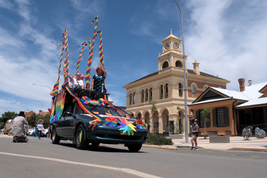 A drag queen in a sparkly dress stands on top of a modified car with other people and rainbow poles.